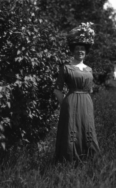 Montgomery modelling an elaborate Edwardian hat and brown poplin dress with button detailing, lace inset sleeves and collar, and front pleats.